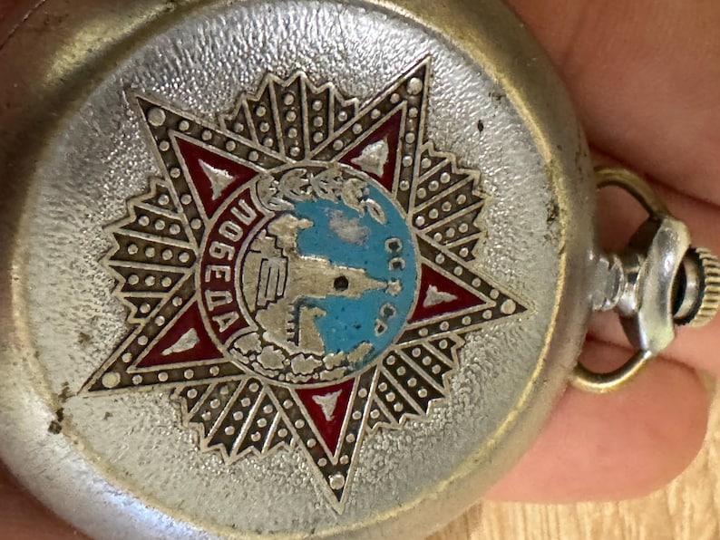 Puede incluir: Reloj de bolsillo plateado con un emblema en forma de estrella que presenta un globo terr&aacute;queo y la palabra "Победа" en letras rojas. El reloj tiene una caja plateada y una tija.