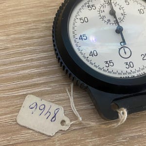 Pu&ograve; includere: Un cronometro nero con quadrante bianco, con numeri e segni neri. Il cronometro ha una lunetta strutturata e una corona di carica. &Egrave; attaccata una piccola etichetta con il numero "8466". Il cronometro &egrave; su una superficie di legno marrone chiaro.