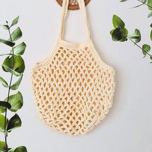 May include: A cream-colored crocheted mesh bag with a woven wood handle hanging on a white wall.