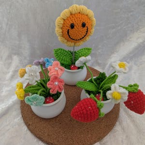 Puede incluir: Tres macetas de flores de ganchillo con un girasol amarillo sonriente, una flor blanca con centro amarillo y una fresa roja. Las macetas son blancas y se encuentran sobre una superficie de corcho.