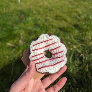 May include: A crocheted donut with white frosting and red stripes. The donut is held in a hand against a green grassy background.
