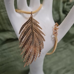 Könnte beinhalten: Eine goldene Feder-Anhänger-Kette mit einem Wildlederband. Die Feder ist mit einem strukturierten Design verziert.