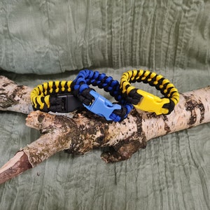 Könnte beinhalten: Drei Paracord-Armbänder mit schwarzen, gelben und blauen gewebten Mustern. Jedes Armband hat einen Plastikverschluss.
