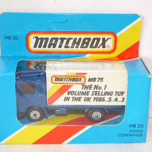 Könnte beinhalten: Ein blauer Matchbox MB 20 Volvo Container-LKW Spielzeug mit einem weißen Kastenanhänger. Die Kiste hat rote und gelbe Streifen und den Text "Matchbox MB 75 The No. 1 Volume Selling Toy in the UK 1986...5...4...3".