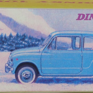 May include: A blue Dinky Toys Fiat 600D toy car with a white roof and black trim. The car is on a snowy background with two people in the background.