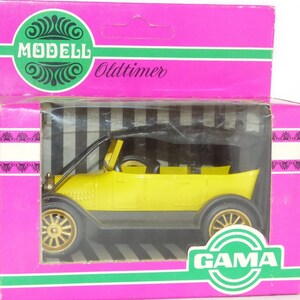 Może przedstawiać: Żółty plastikowy samochód z czarnym dachem i złotymi kołami. Samochód znajduje się w plastikowym pudełku z czarno-białym paskowanym tłem. Na pudełku widnieją napisy "Modell Oldtimer" i "GAMA".