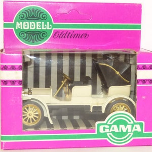 Puede incluir: Un modelo de coche antiguo blanco en una caja rosa con una etiqueta verde y blanca que dice "Modell Oldtimer" y "GAMA".