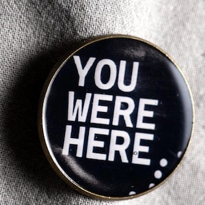 May include: A circular pin badge with a black background and white text that reads YOU WERE HERE. The badge features a thin metallic gold-toned rim and a smooth, glossy finish, with three small white dots visible in the lower right corner.