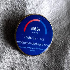 Könnte beinhalten: Ein runder Ansteckpin mit dunkelblauer Oberfläche, der eine rot-blaue Bogenanzeige mit 56 % zeigt. Der Text lautet: 56% High risk, High risk — not recommended right now, more people are trying to catch the perfect window. Der Pin hat einen Goldrand.
