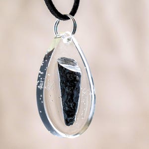 May include: A teardrop-shaped pendant made of clear resin containing a jagged, dark black mineral fragment. The pendant is suspended from a black cord by a silver-toned metal jump ring, with small air bubbles visible within the resin material.