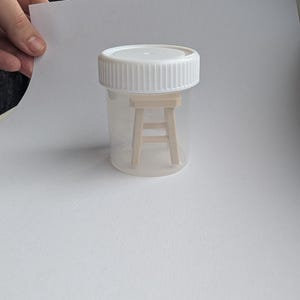 Peut inclure: Un r&eacute;cipient en plastique transparent avec un couvercle blanc contenant un tabouret en bois miniature. Le tabouret est brun clair et poss&egrave;de quatre pieds. Le r&eacute;cipient est tenu par une main sur fond blanc.