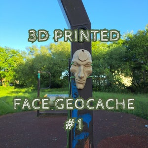 May include: A beige 3D printed face geocache container attached to a dark metal pole with green string. The text 3D PRINTED FACE GEOCACHE # 1 is overlaid on the image. The face features a hexagonal shape on the forehead and a small hole at the top.