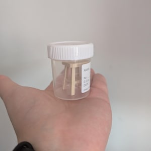 Peut inclure: Un petit tabouret en bois &agrave; l'int&eacute;rieur d'un r&eacute;cipient en plastique transparent avec un couvercle blanc. Le r&eacute;cipient porte une &eacute;tiquette avec les mots "NAME", "No.", "D.O.B", "DATE" et "SINGLE".