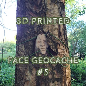 May include: A 3D printed beige face mask geocache container with a hexagonal detail on the forehead. The mask is secured to a tree trunk with thin green cord threaded through holes in the mask. The text 3D PRINTED FACE GEOCACHE # 5 is visible.