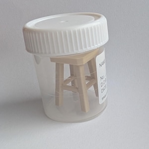 Peut inclure: Un petit tabouret en bois &agrave; l'int&eacute;rieur d'un r&eacute;cipient en plastique transparent avec un couvercle blanc. Le r&eacute;cipient porte une &eacute;tiquette avec du texte. Le tabouret est brun clair et a quatre pieds.