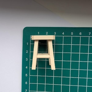 Peut inclure: Un petit tabouret en bois non fini, avec une assise rectangulaire et quatre pieds inclin&eacute;s. Il est pos&eacute; sur un tapis de d&eacute;coupe vert avec des lignes de quadrillage et des num&eacute;ros. Il s'agit probablement d'une maquette.
