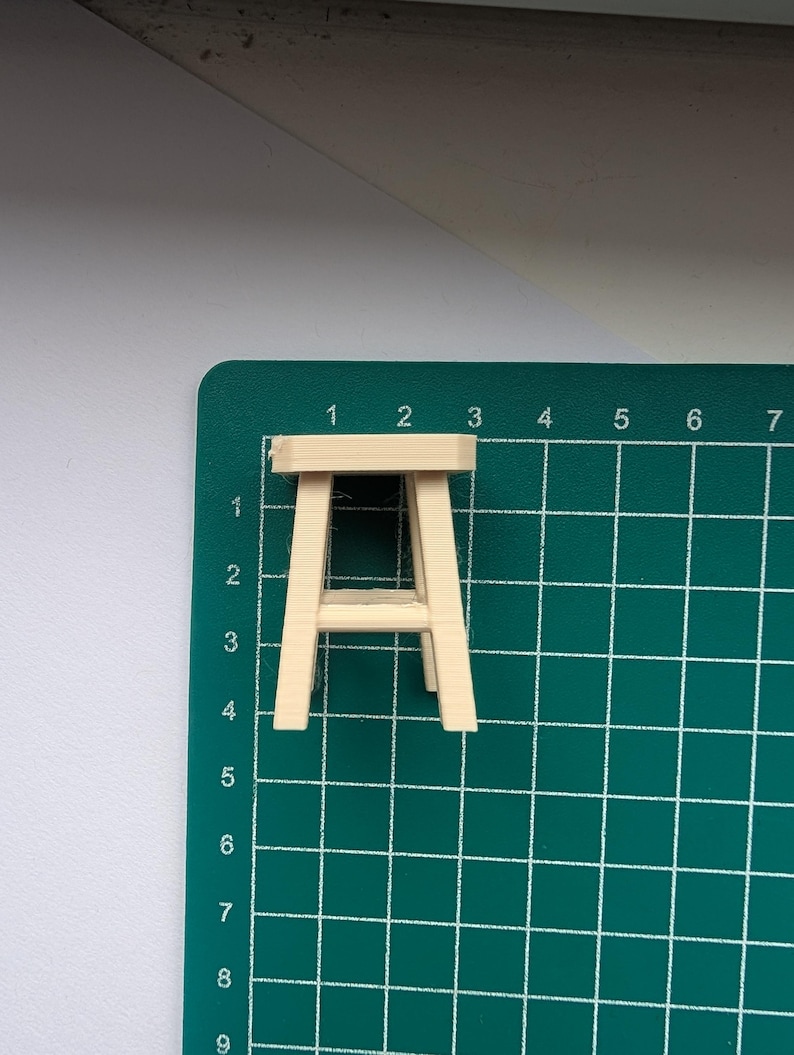 Peut inclure: Un petit tabouret en bois non fini, avec une assise rectangulaire et quatre pieds inclin&eacute;s. Il est pos&eacute; sur un tapis de d&eacute;coupe vert avec des lignes de quadrillage et des num&eacute;ros. Il s'agit probablement d'une maquette.
