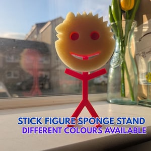 May include: A novelty sponge holder in the shape of a stick figure. The sponge is yellow with red eyes and a red smile. The stick figure stand is red. Text on the image reads "STICK FIGURE SPONGE STAND DIFFERENT COLOURS AVAILABLE."