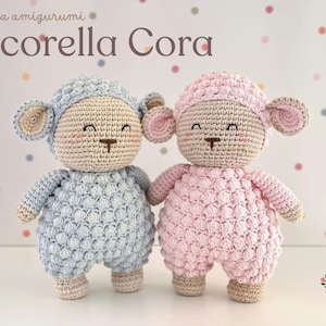 May include: Two handmade crochet sheep dolls, one light blue and one pink, stand side-by-side. Each doll has a textured body, a matching bonnet, and beige accents. The dolls have smiling faces and are labeled "Pecorella Cora".