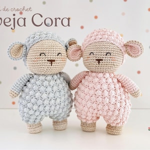 May include: Two handmade crochet sheep dolls, one light blue and one pink, stand side-by-side. Each doll has a textured body, a matching hat, and beige ears. The dolls have smiling faces with brown stitched details. The text "Oveja Cora" is above them.