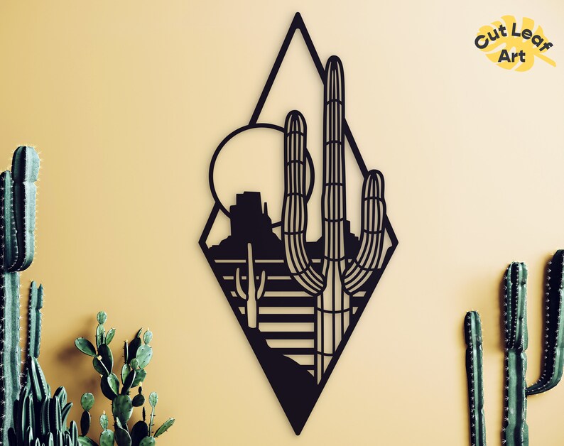 May include: Black metal wall art featuring a desert landscape with a cactus and a mountain in a diamond shape. The art is on a light yellow background.
