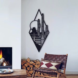 May include: Black metal wall art in the shape of a diamond featuring a silhouette of a desert landscape with a cactus, mountains, and a sun.