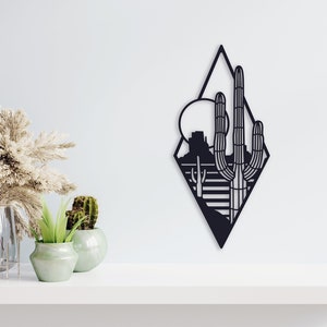 May include: Black metal wall art featuring a desert landscape with a large cactus and a mountain in the background. The art is in the shape of a diamond.