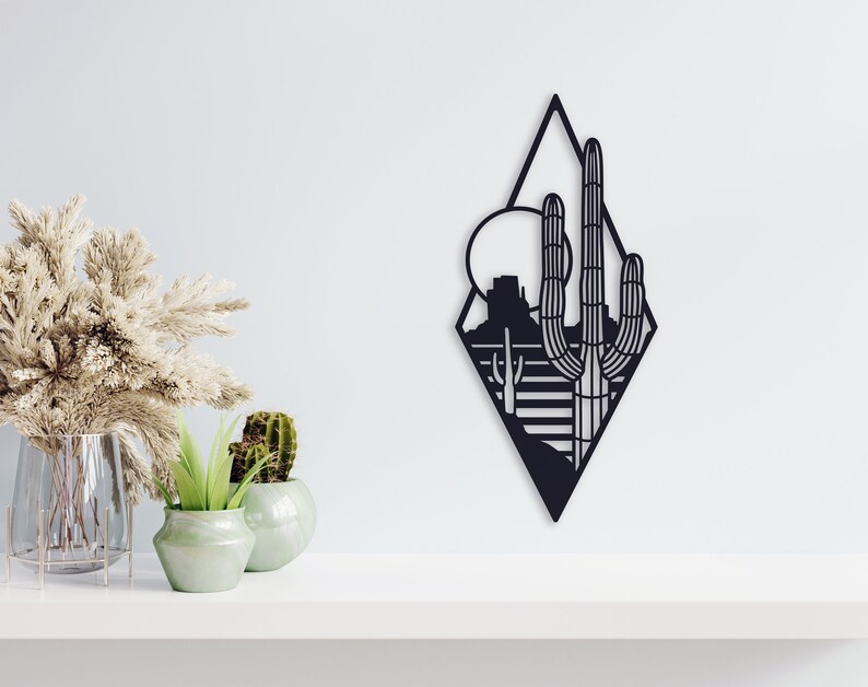 May include: Black metal wall art featuring a desert landscape with a large cactus and a mountain in the background. The art is in the shape of a diamond.