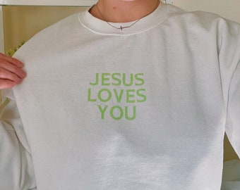 May include: White sweatshirt with the text "JESUS LOVES YOU" in green. The sweatshirt is a crew neck style and appears to be made of a soft material. The text is centered on the front of the sweatshirt.