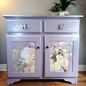 May include: A lavender-coloured cabinet with two drawers and two doors. The doors feature a floral print and the drawers have silver butterfly handles.