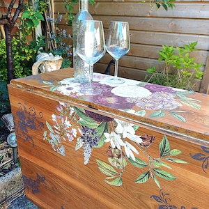 May include: A wooden table with a floral design, holding a clear wine bottle and two wine glasses. The table has a warm brown tone with a detailed botanical pattern in purple, white, and green. The scene is set outdoors with greenery in the background.