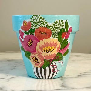 Puede incluir: Una maceta de terracota azul claro con un diseño pintado de un ramo de flores rosas, rojas y amarillas en un jarrón de rayas blancas y negras.