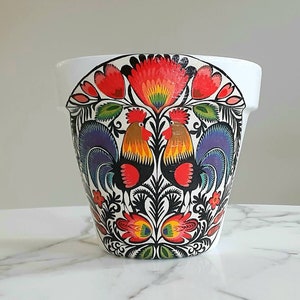 May include: White ceramic planter with a folk art design featuring two roosters with red, yellow, blue, and green accents. The design is on a white background.