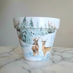 Puede incluir: Maceta de cerámica blanca con un diseño de escena de invierno. La maceta presenta dos ciervos en un bosque nevado con árboles y una pequeña cabaña en el fondo.