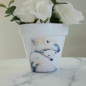 May include: White ceramic flower pot featuring a painted design of three polar bears. The pot is filled with white roses and green leaves.