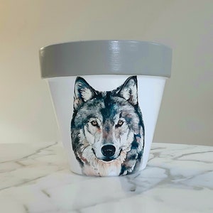 May include: A white ceramic planter with a gray rim features a watercolor painting of a wolf's face. The wolf has a gray and black coat and a serious expression.