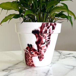 May include: A white ceramic planter featuring a painted design of two giraffes, one with its head tilted down to nuzzle the other. The planter is filled with a green leafy plant.