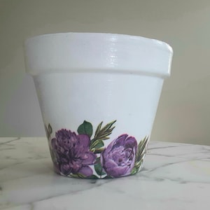 Puede incluir: Maceta de cerámica blanca con un diseño floral morado alrededor de la base.
