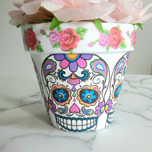May include: White ceramic flower pot with a colorful Day of the Dead sugar skull design. The pot is decorated with pink roses around the top rim.