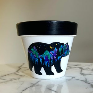 May include: A white terracotta planter with a black rim, featuring a hand-painted design of a bear with a mountain range and starry night sky inside its silhouette.