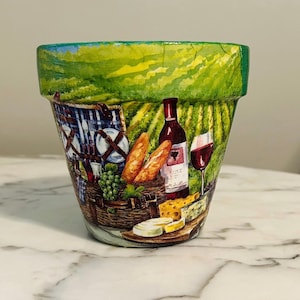 May include: A hand-painted terracotta flower pot featuring a picnic scene. The design includes a picnic basket, wine bottle, wine glass, bread, cheese, and grapes. The background depicts a vineyard with green and yellow hues.