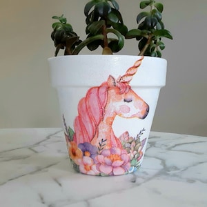 Könnte beinhalten: Weißer Keramik-Blumentopf mit handgemaltem Einhorn-Design. Das Einhorn hat eine rosa Mähne und ein goldenes Horn, umgeben von bunten Blumen. Der Topf ist mit grünen Sukkulenten gefüllt.