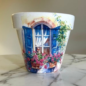 May include: White ceramic flower pot with a painted design of a blue window with white curtains and pink flowers in pots on a windowsill.