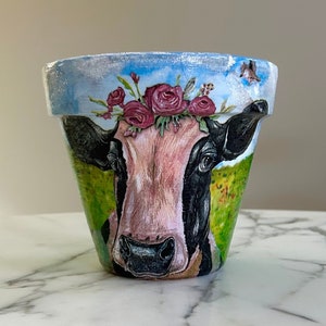 May include: A white ceramic flower pot with a painted image of a cow wearing a flower crown. The cow is looking directly at the viewer. The background is a green field with a blue sky.