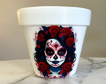 Puede incluir: Maceta de cerámica blanca con un diseño colorido de calavera de azúcar del Día de Muertos con rosas rojas.