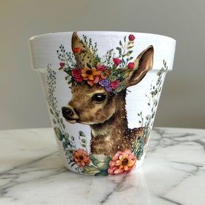 Puede incluir: Maceta de cerámica blanca con una ilustración detallada de la cabeza de un ciervo adornada con una corona floral. El ciervo es marrón con un fondo blanco y flores coloridas. Un artículo decorativo para el hogar o el jardín.