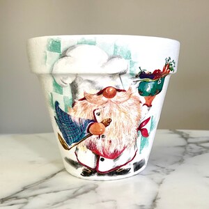May include: White ceramic planter with a hand-painted design of a chef gnome holding a whisk and a bowl of vegetables. The gnome is wearing a white chef hat, a red and white striped shirt, and a blue and green plaid apron.