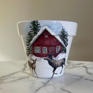 May include: White ceramic planter with a winter scene design. The design features a red cabin with a snowy roof, two deer, and pine trees.