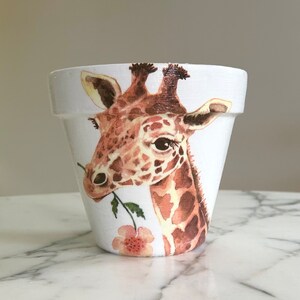 Puede incluir: Maceta de cerámica blanca con una imagen pintada de una jirafa comiendo una flor. La jirafa es marrón y blanca con una flor rosa.