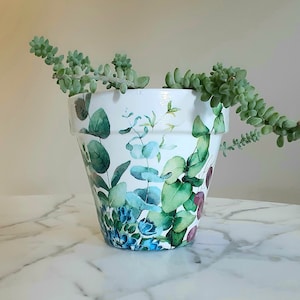 May include: White ceramic planter with a floral design featuring green leaves and blue flowers. The planter is filled with a succulent plant.